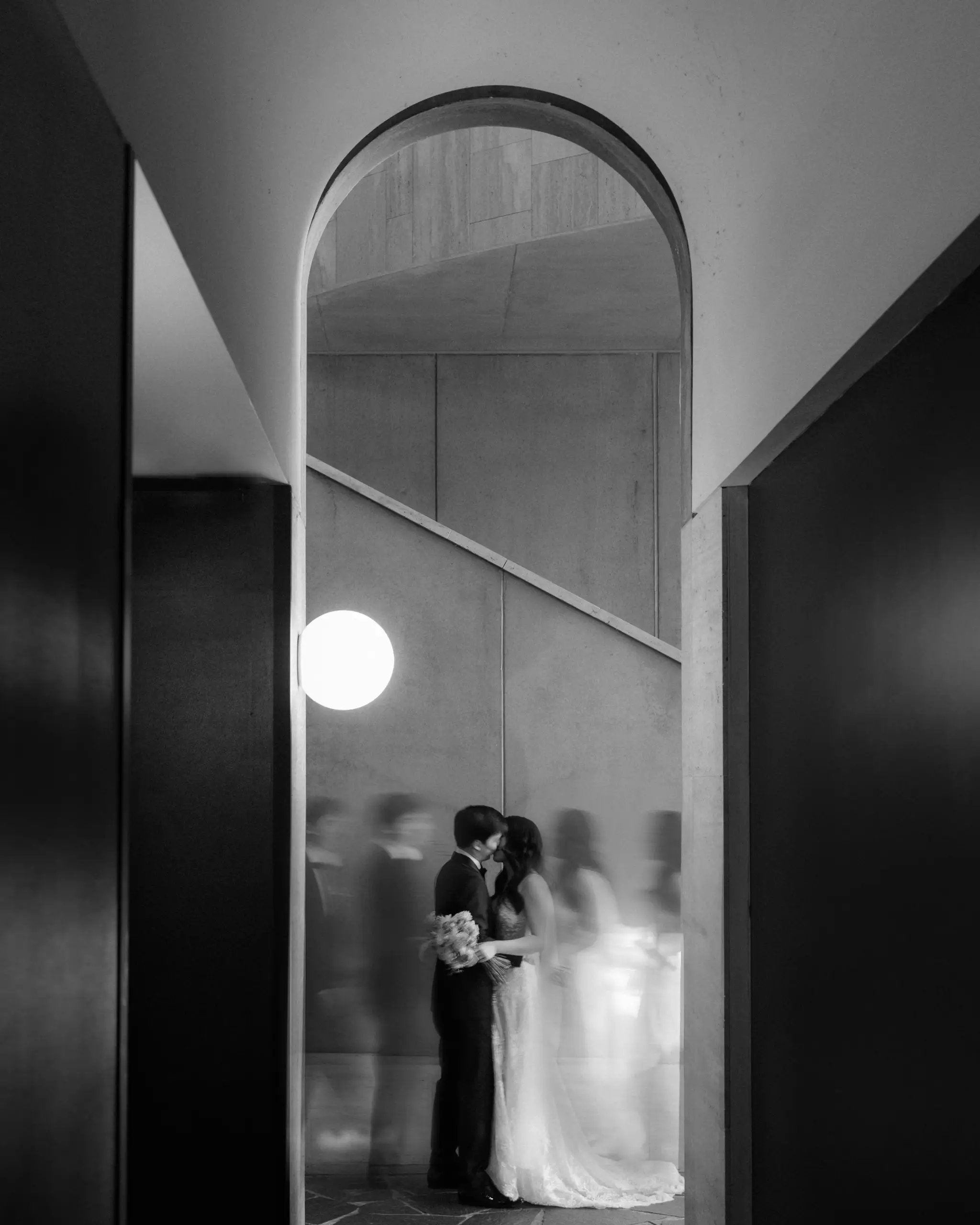 The couple kissing beneath the concrete arch, a globe lamp glowing beside them and guests blurring past, The Calile Hotel, Brisbane
