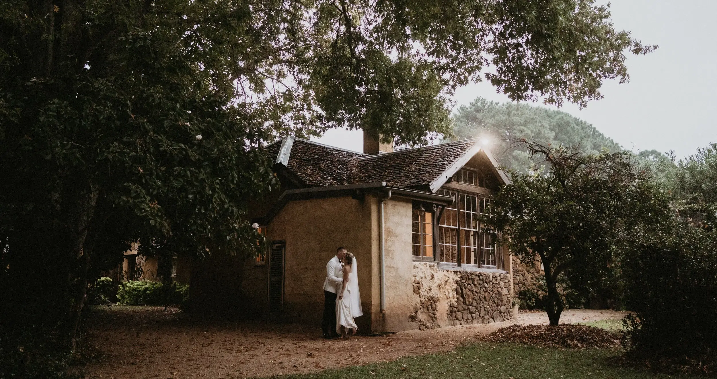Maddie and John embracing beside the old stone cottage in the garden at Deux Belettes, Byron Bay hinterland