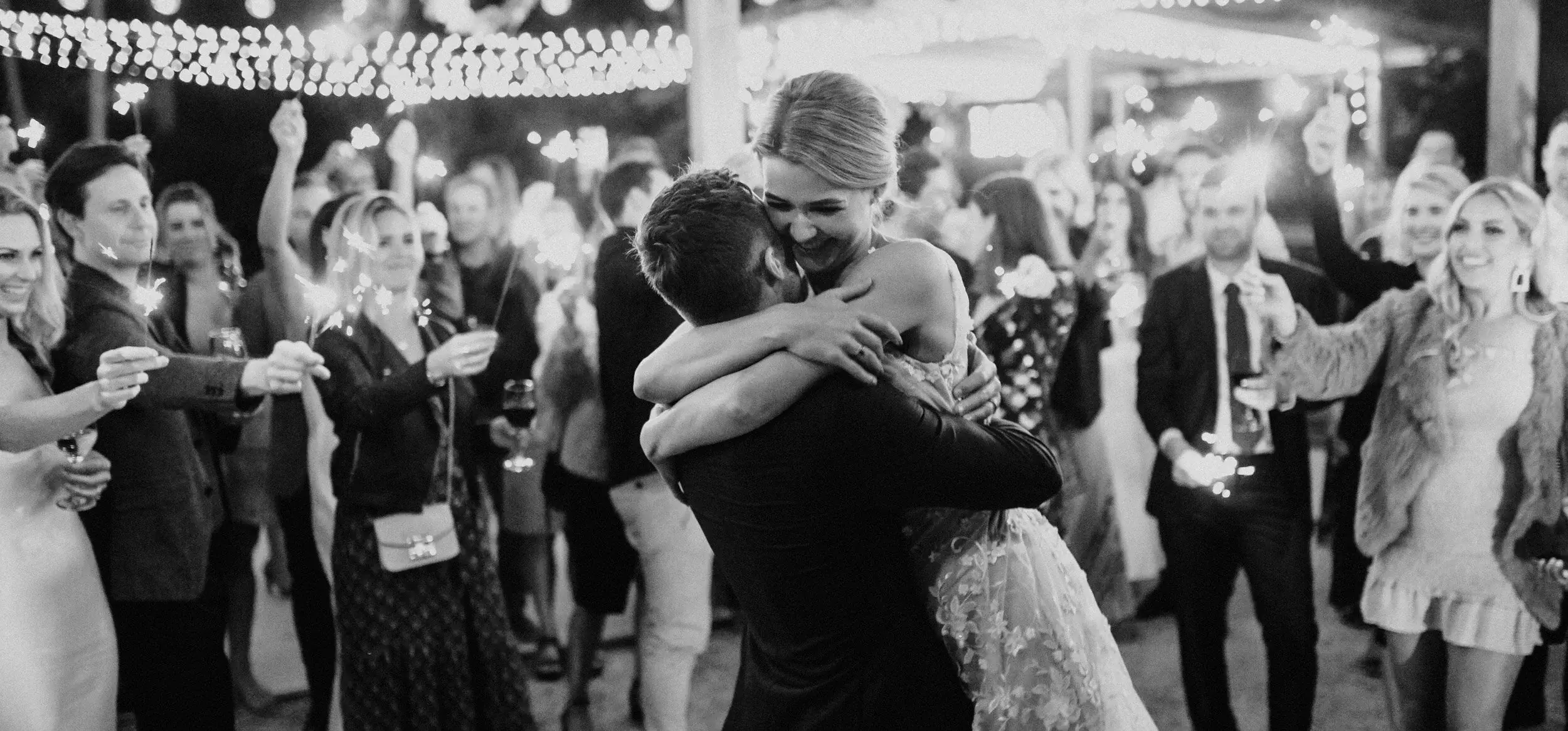 Em and Nev holding each other on the dance floor at night, guests circling them with sparklers under strings of fairy lights