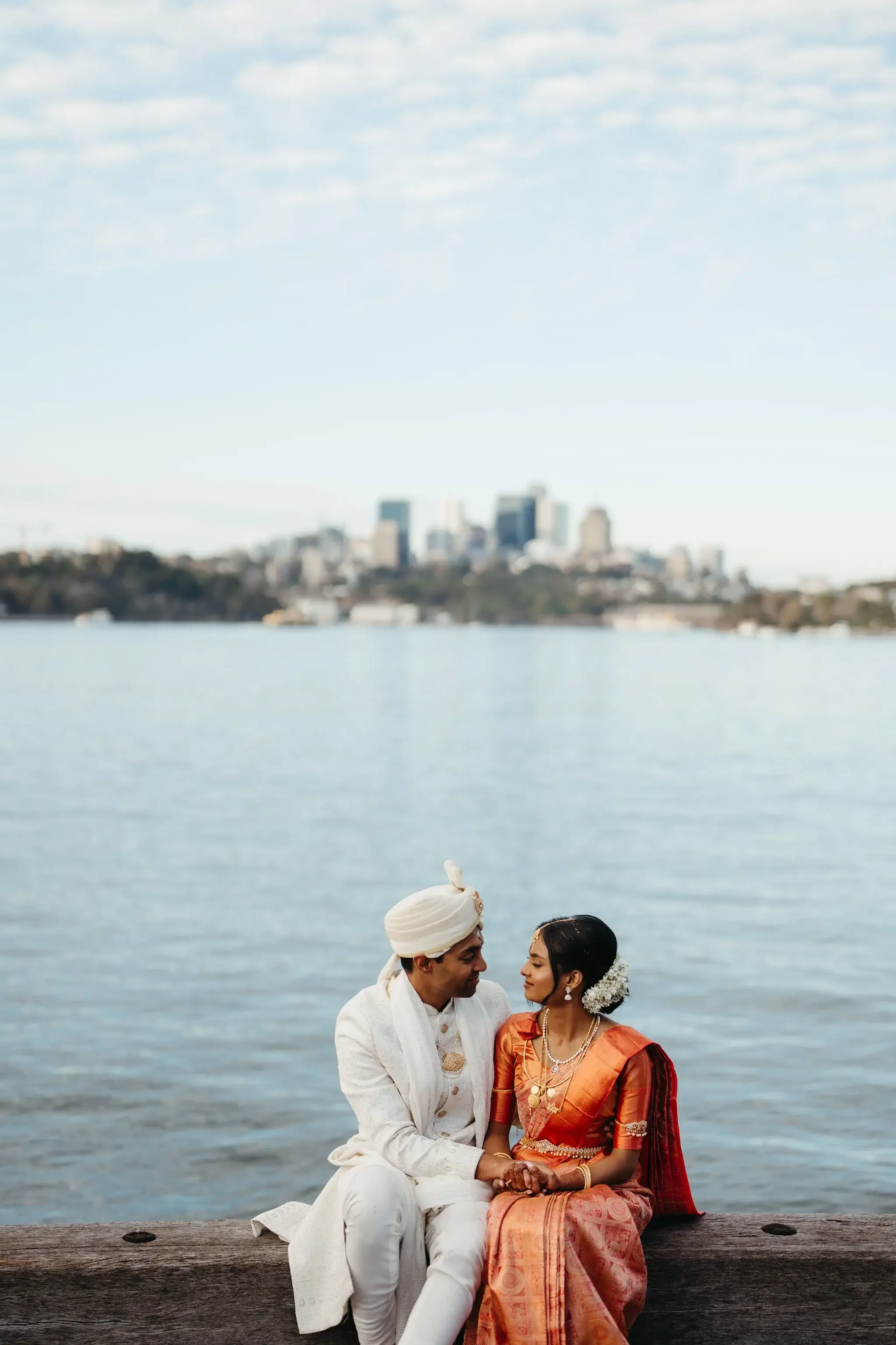 A couple in white and red traditional dress seated on rocks above Sydney Harbour, city skyline behind