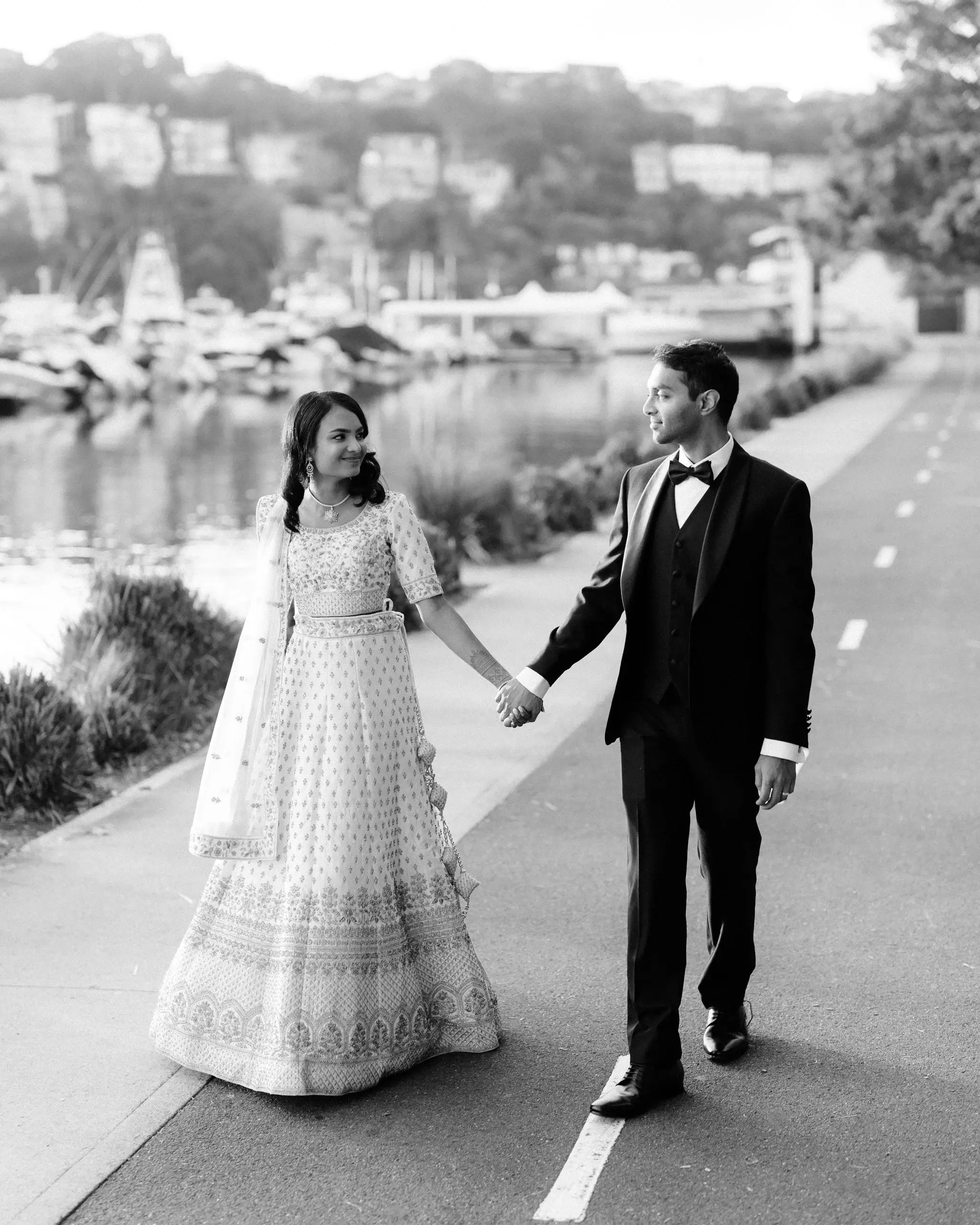 Black and white photograph of a couple holding hands on a harbourside path, boats moored behind