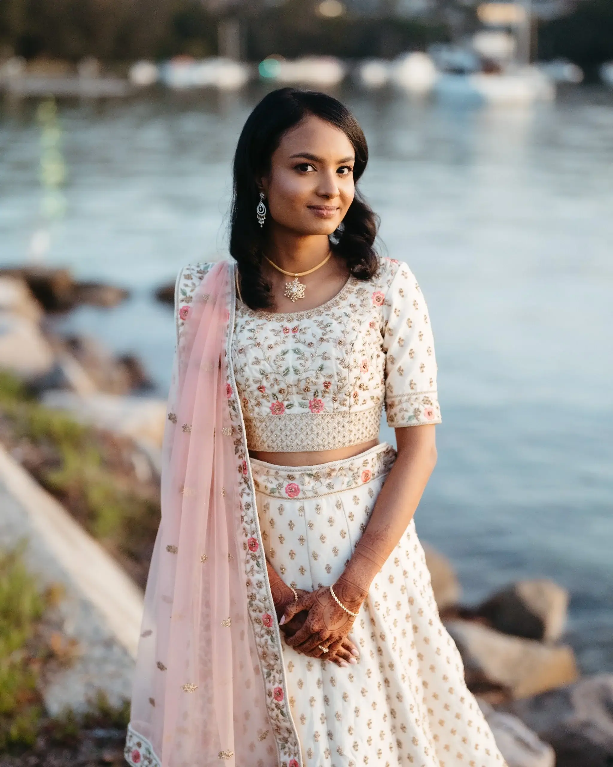Portrait of a bride in a pale embroidered lehenga and pink dupatta beside the water