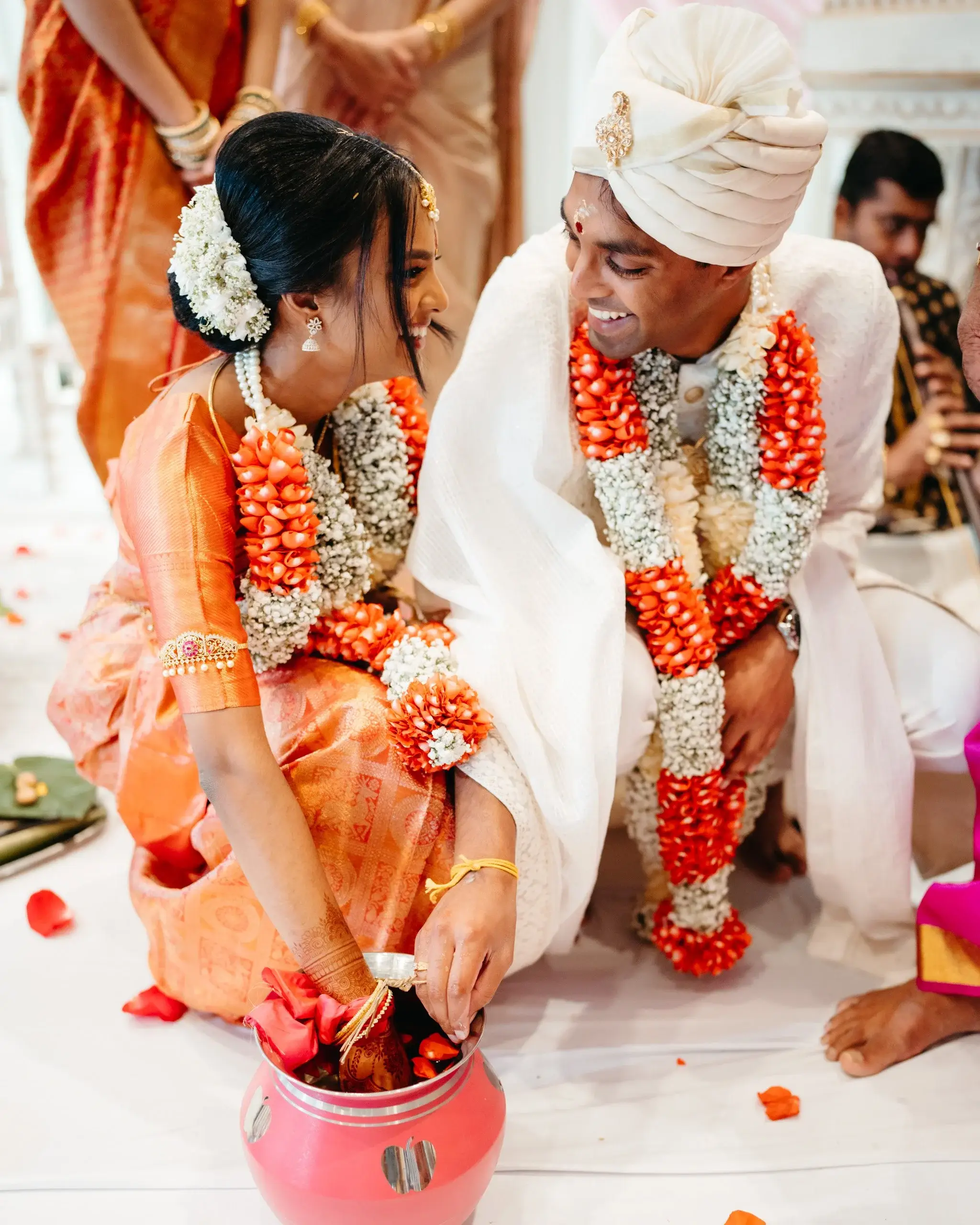 A couple in orange and gold traditional dress performing a ritual over a decorated pot